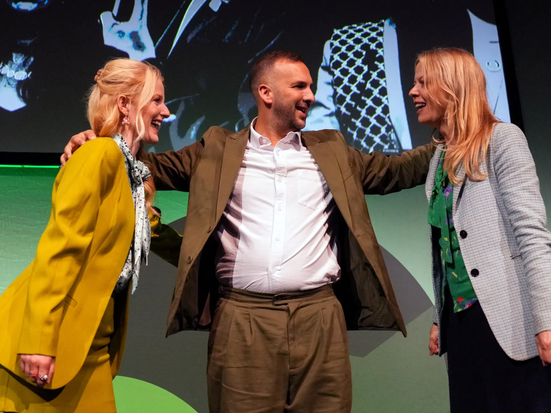 A man stands between two women onstage, smiling with his arms around their shoulders.