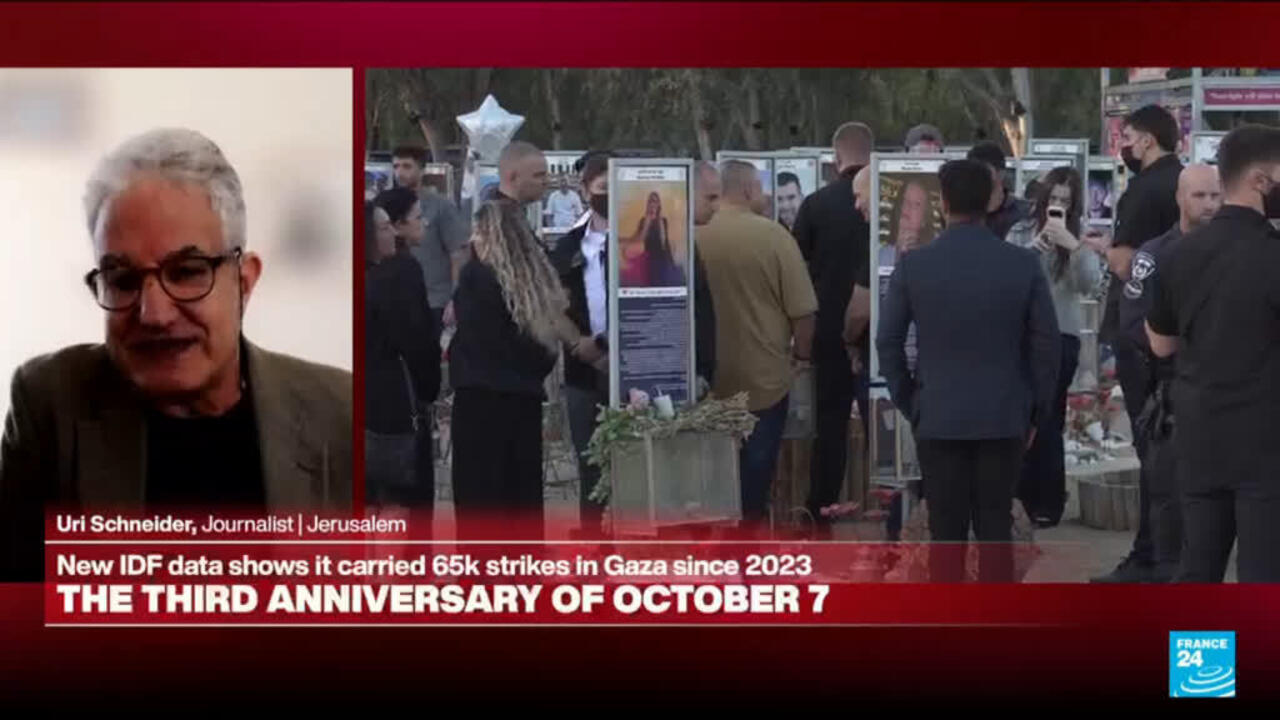 A man speaks in a broadcast panel beside crowds viewing memorial displays and photographs outdoors.