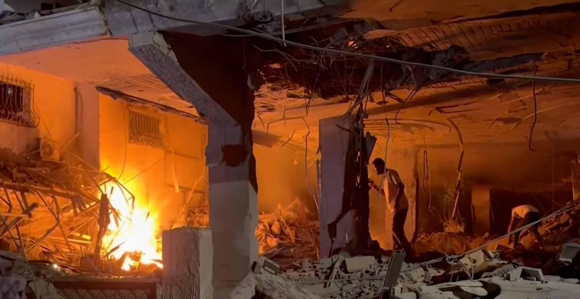 Fire burns amid rubble beneath a collapsed building, as two people move through the wreckage.