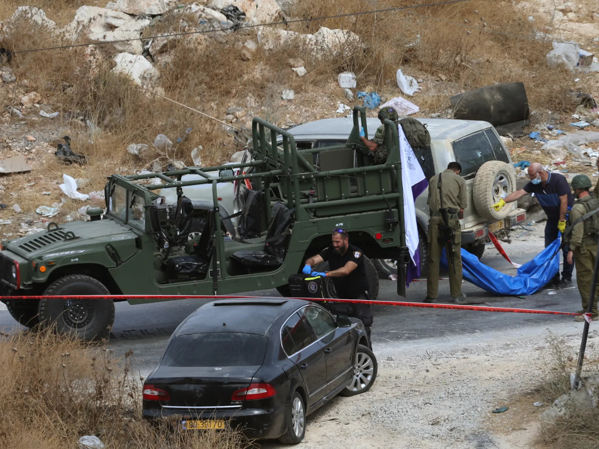 Israeli forces and emergency personnel gather beside vehicles as a blue-covered stretcher is handled at a roadside scene.