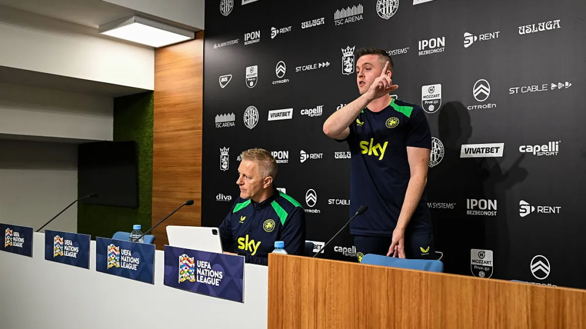 A standing man gestures beside a seated man at a football news conference table.