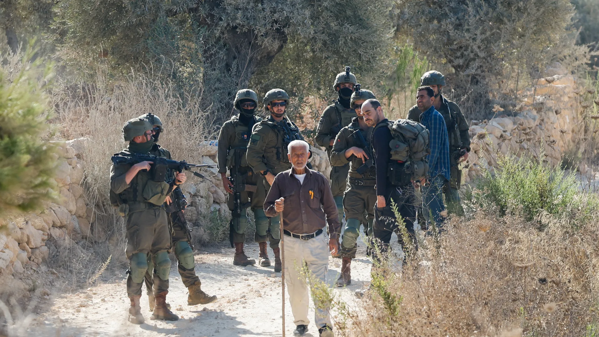 An elderly man with a cane walks among armed soldiers and another man on a rural path.