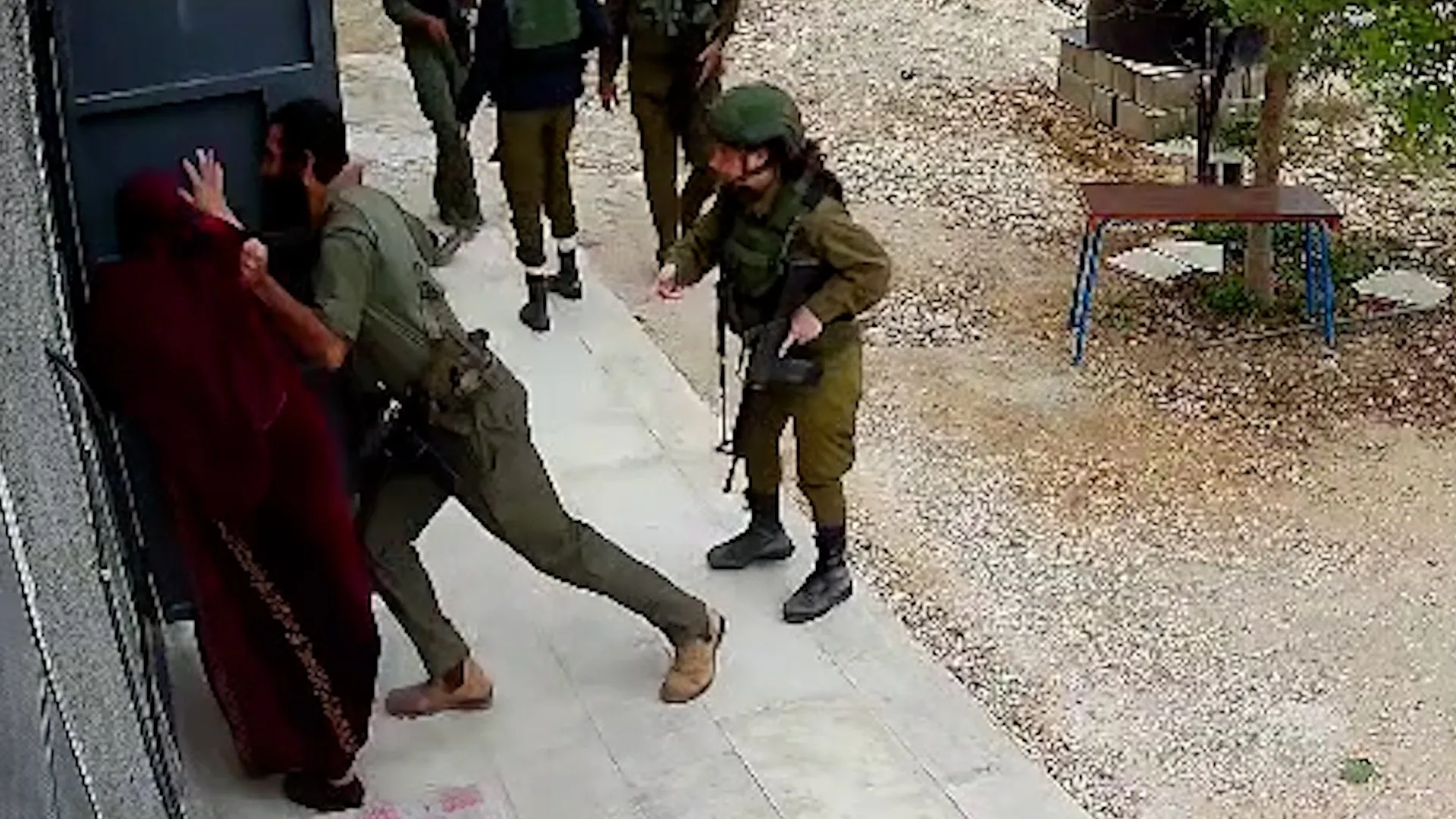 A uniformed soldier appears to kick a woman in a burgundy robe beside a doorway as others stand nearby.