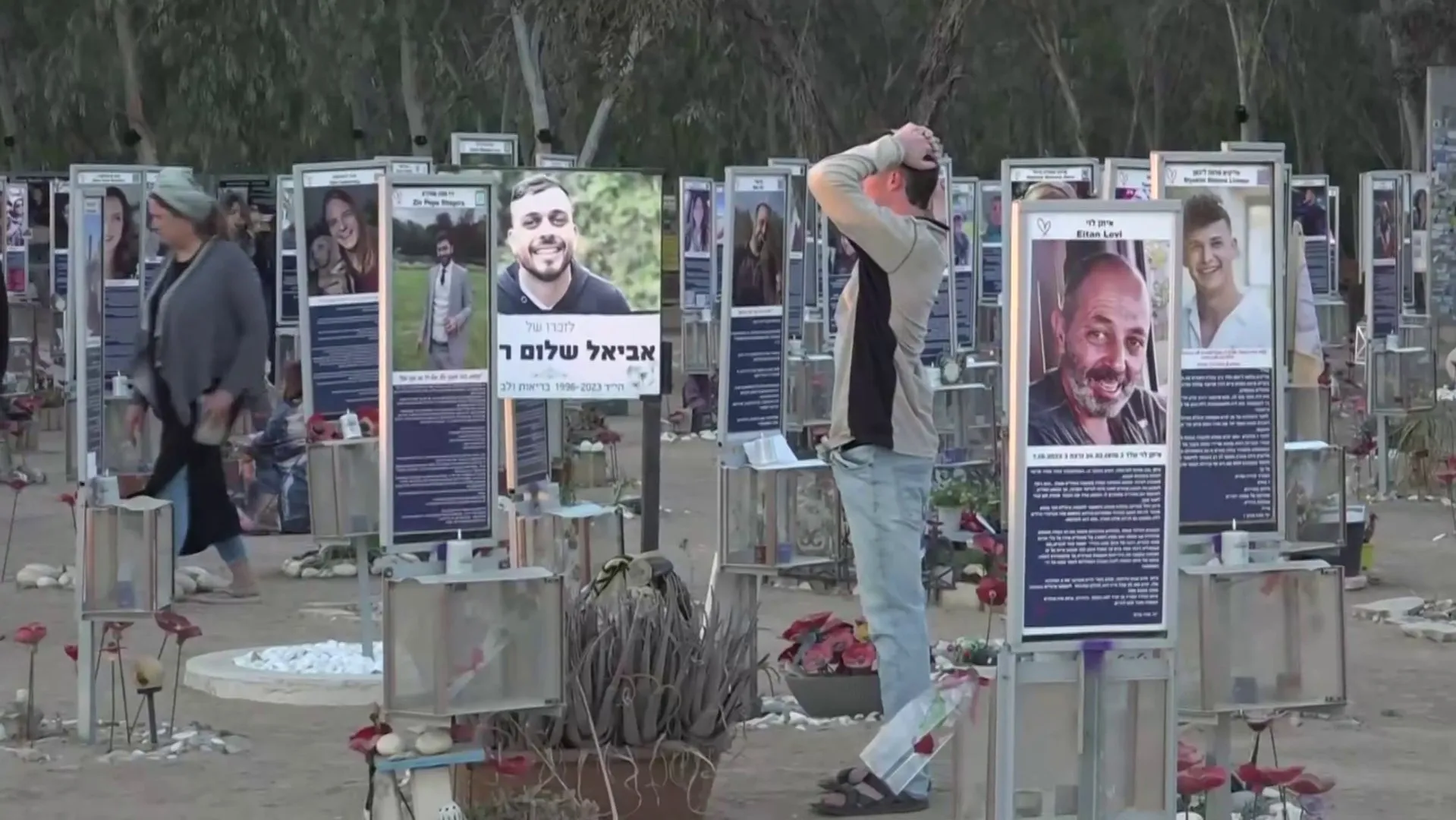 People walk among memorial portrait displays, flowers, and candles at an outdoor remembrance site.