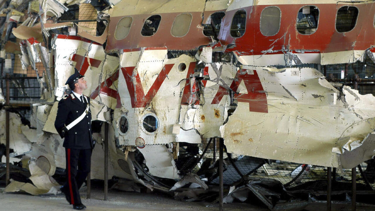 Uniformed officer stands beside wreckage of an aircraft fuselage with red and white markings.