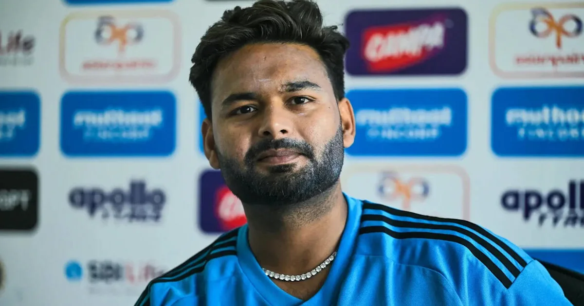 A bearded man in a blue sports jersey sits before a wall of sponsor logos.