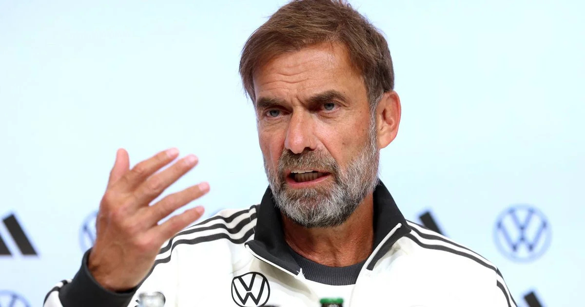 Bearded man in a white Adidas jacket gestures while speaking at a press event.