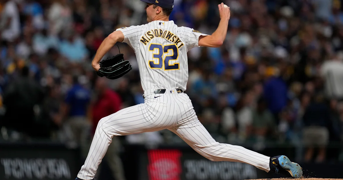 A pitcher in a white pinstriped uniform and number 32 strides forward to throw, with a blurred crowd behind him.