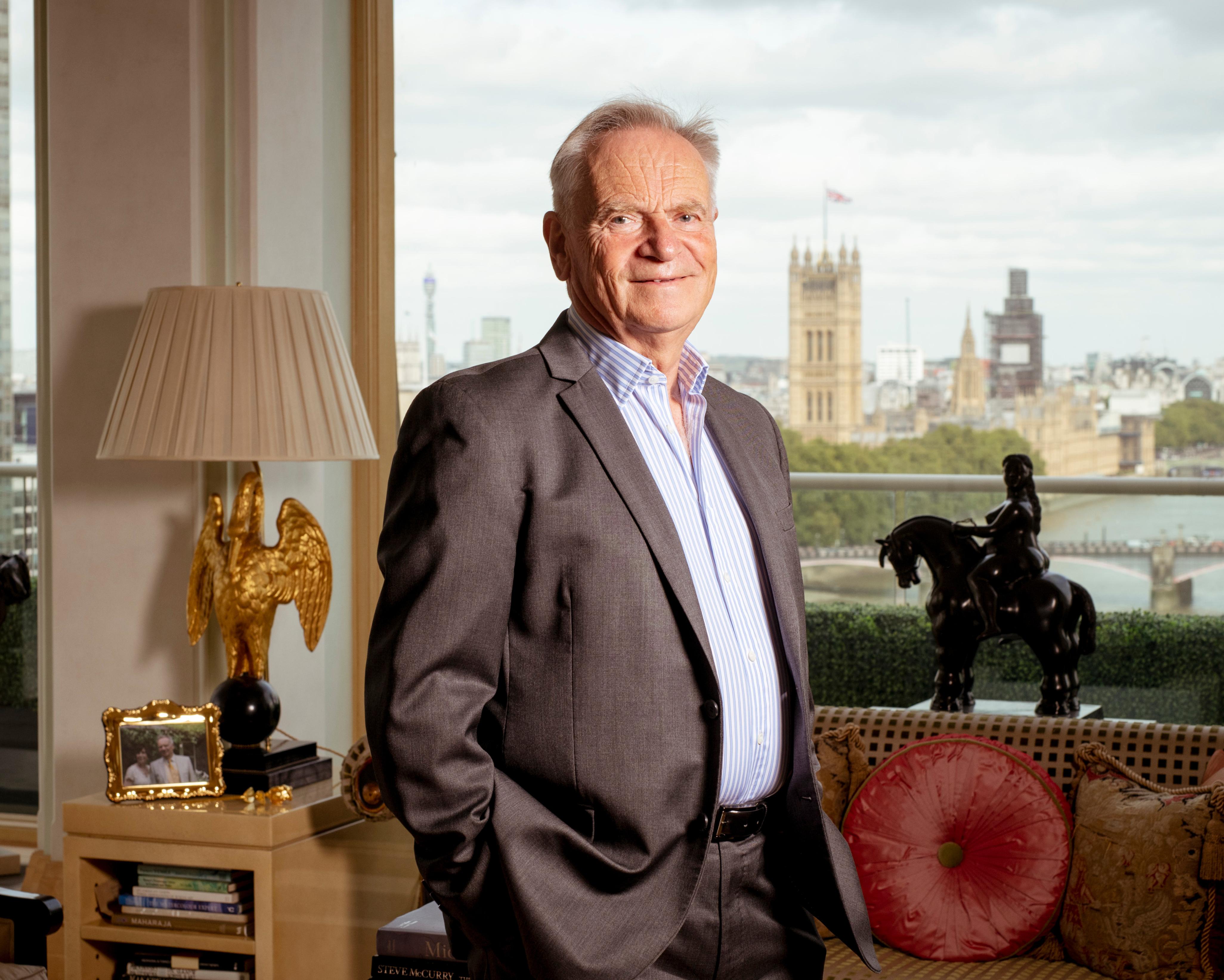 An older man in a suit stands in a furnished room beside a large window overlooking a city skyline.