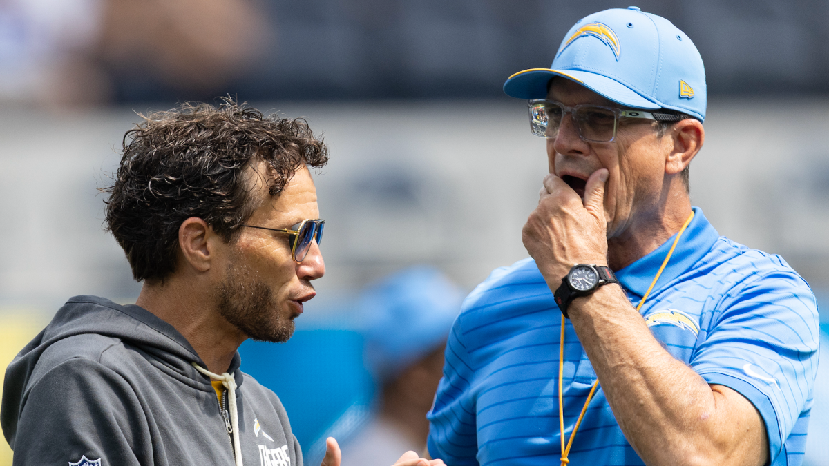 Two men in Chargers apparel talk on the sideline, one wearing sunglasses and the other touching his face.