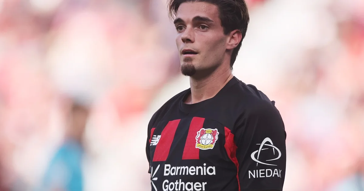 Footballer in a black and red Bayer Leverkusen jersey stands against a blurred stadium background.