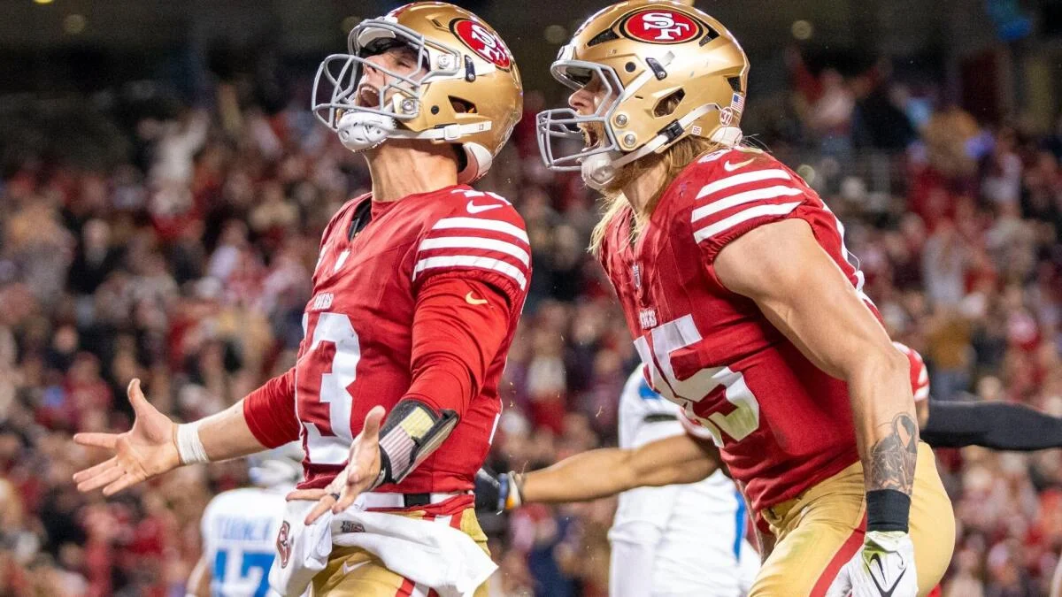 Two San Francisco 49ers players in red jerseys and gold helmets celebrate on the field.