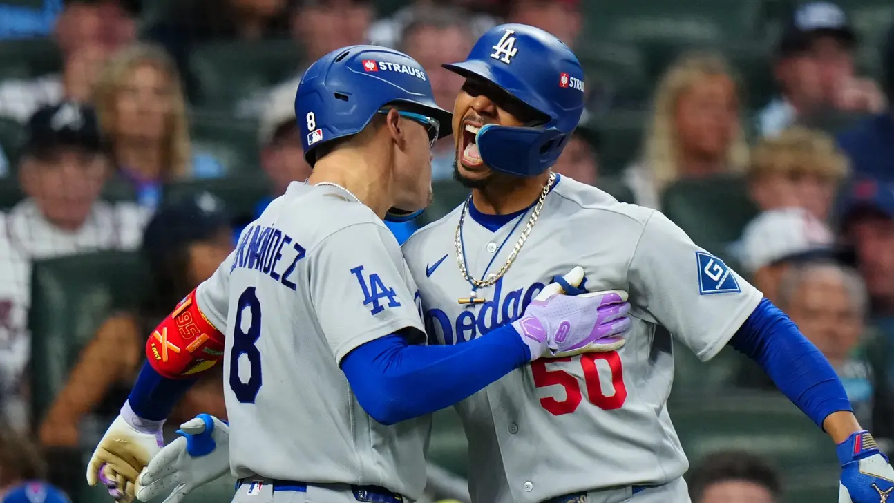 Two Los Angeles Dodgers players in gray uniforms celebrate together on the field, with blurred spectators behind them.