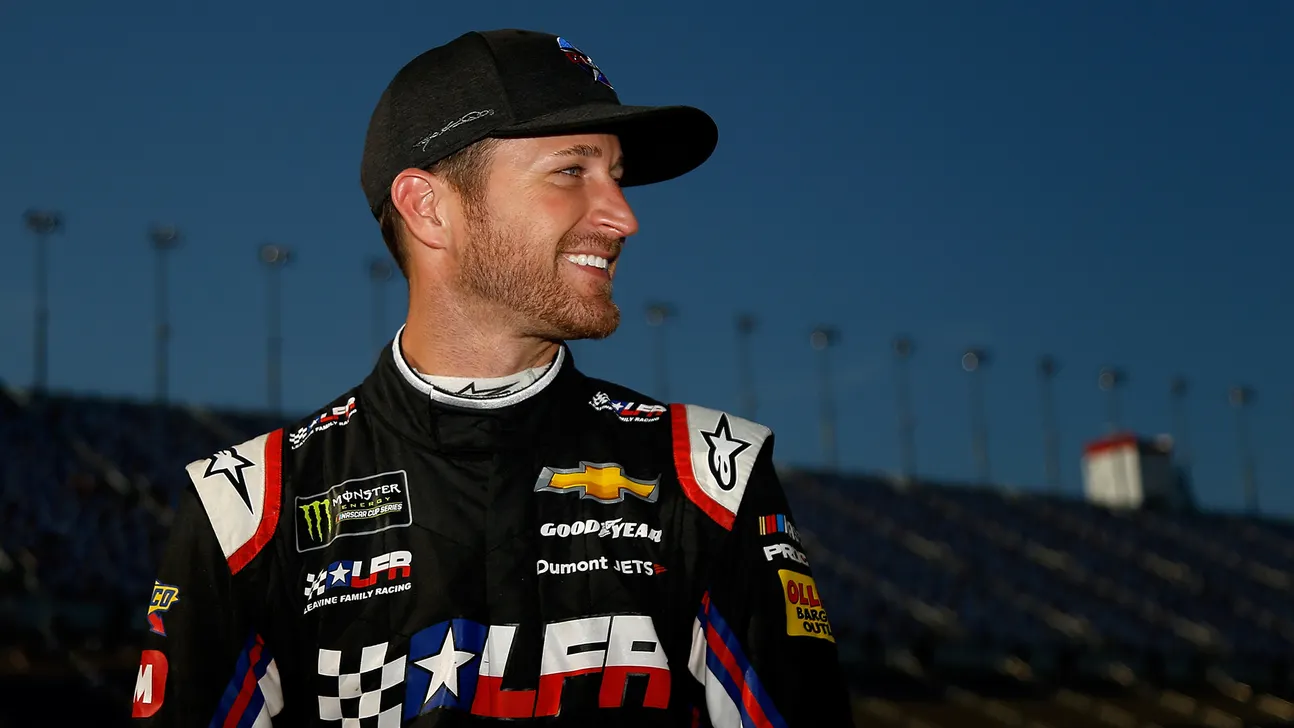 Smiling race car driver in a black sponsor-covered suit and cap stands at a racetrack.