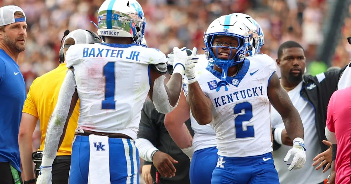 Kentucky players in white and blue uniforms celebrate together on the sideline during a football game.