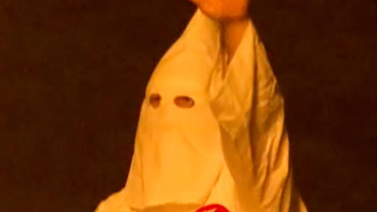 A person in a white hood and robe with eye openings, shown against a dark background.