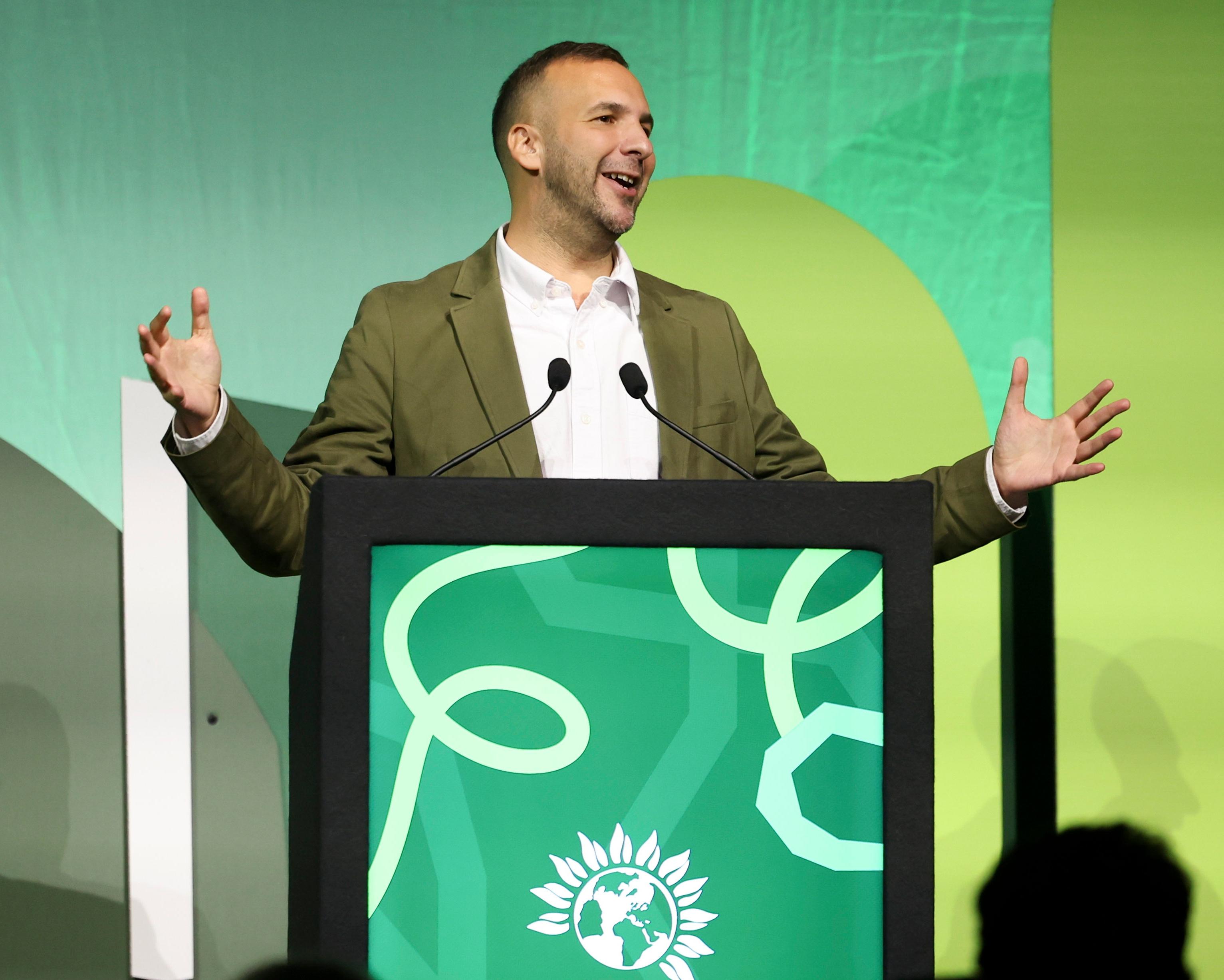 A man in an olive jacket speaks at a lectern with both hands raised against a green backdrop.