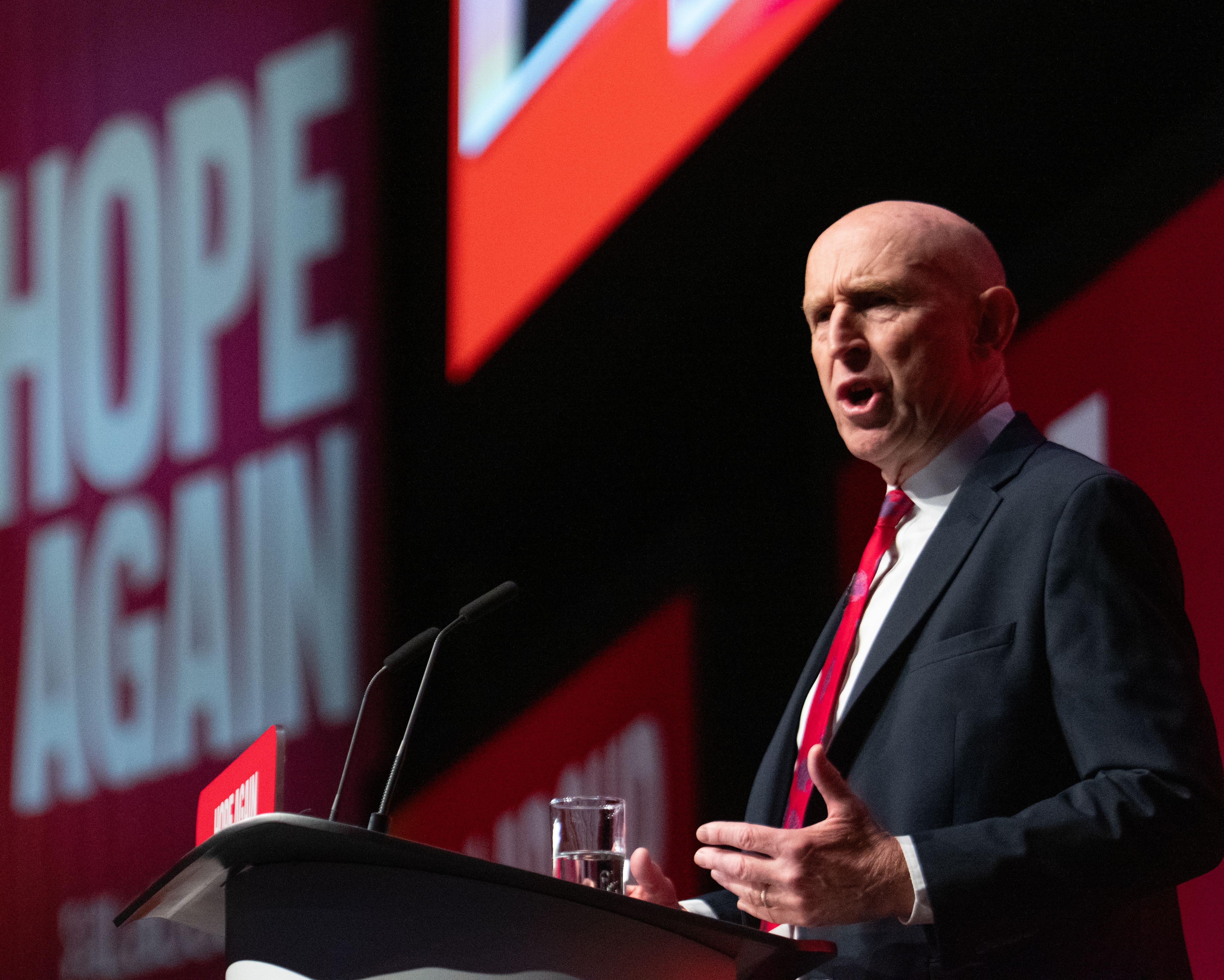 Bald man in a dark suit and red tie speaks at a lectern against a red and black backdrop.