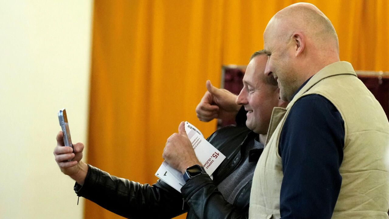 Two men smile while looking at a smartphone; one holds a printed ballot or leaflet.