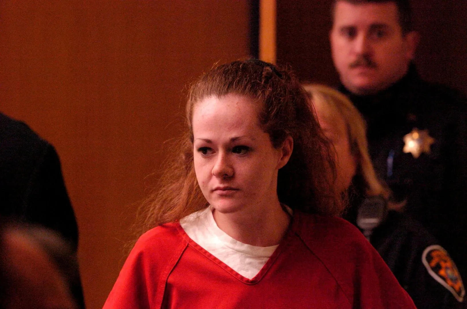 A woman in a red top stands in a courtroom, with uniformed officers behind her.