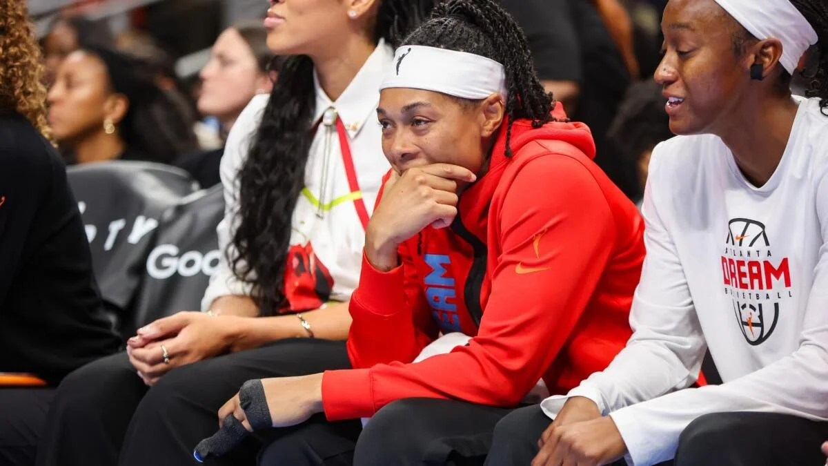 A player in a red hoodie and white headband sits courtside between teammates during a basketball game.