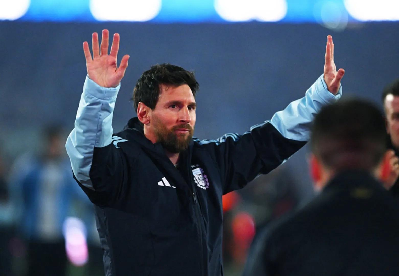 Bearded footballer in a dark jacket raises both hands toward a crowd in a stadium.