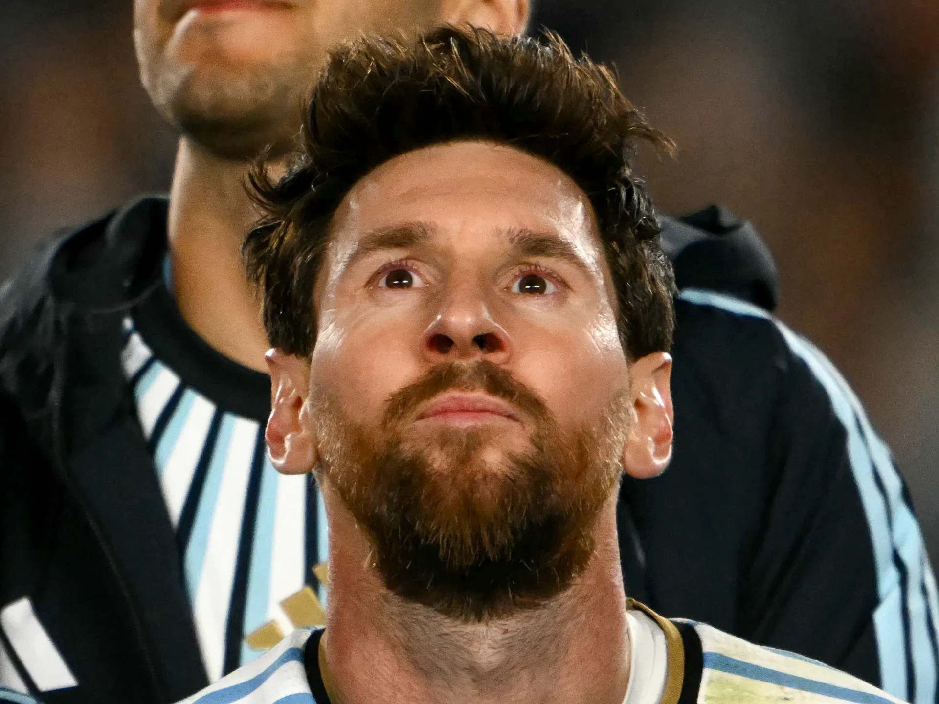 Close-up of a bearded footballer looking upward, with a referee in a striped shirt behind him.