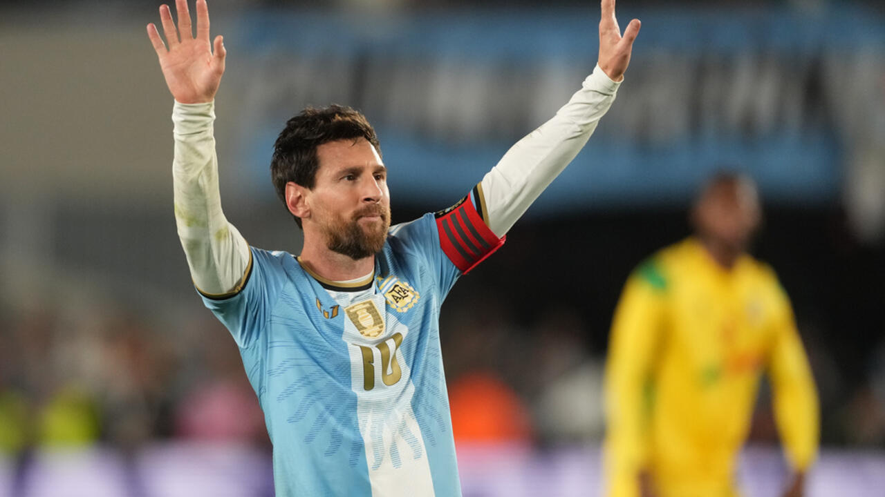 Argentina player in a light blue jersey raises both arms, with a yellow-clad player blurred behind him.