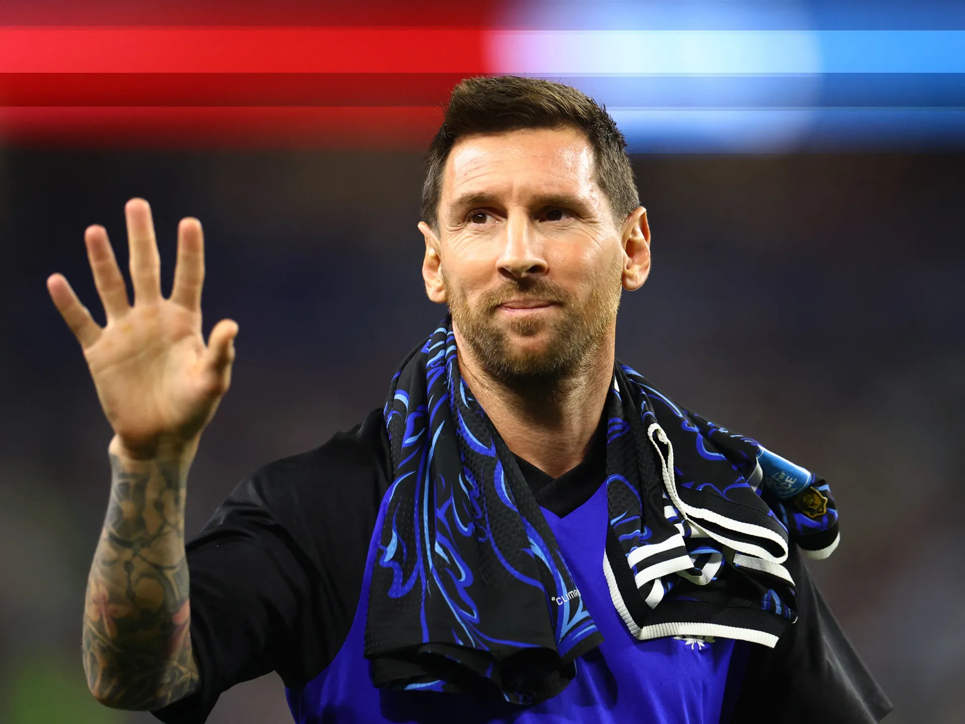 Lionel Messi raises a tattooed hand, wearing a blue and black shirt with scarves around his shoulders.