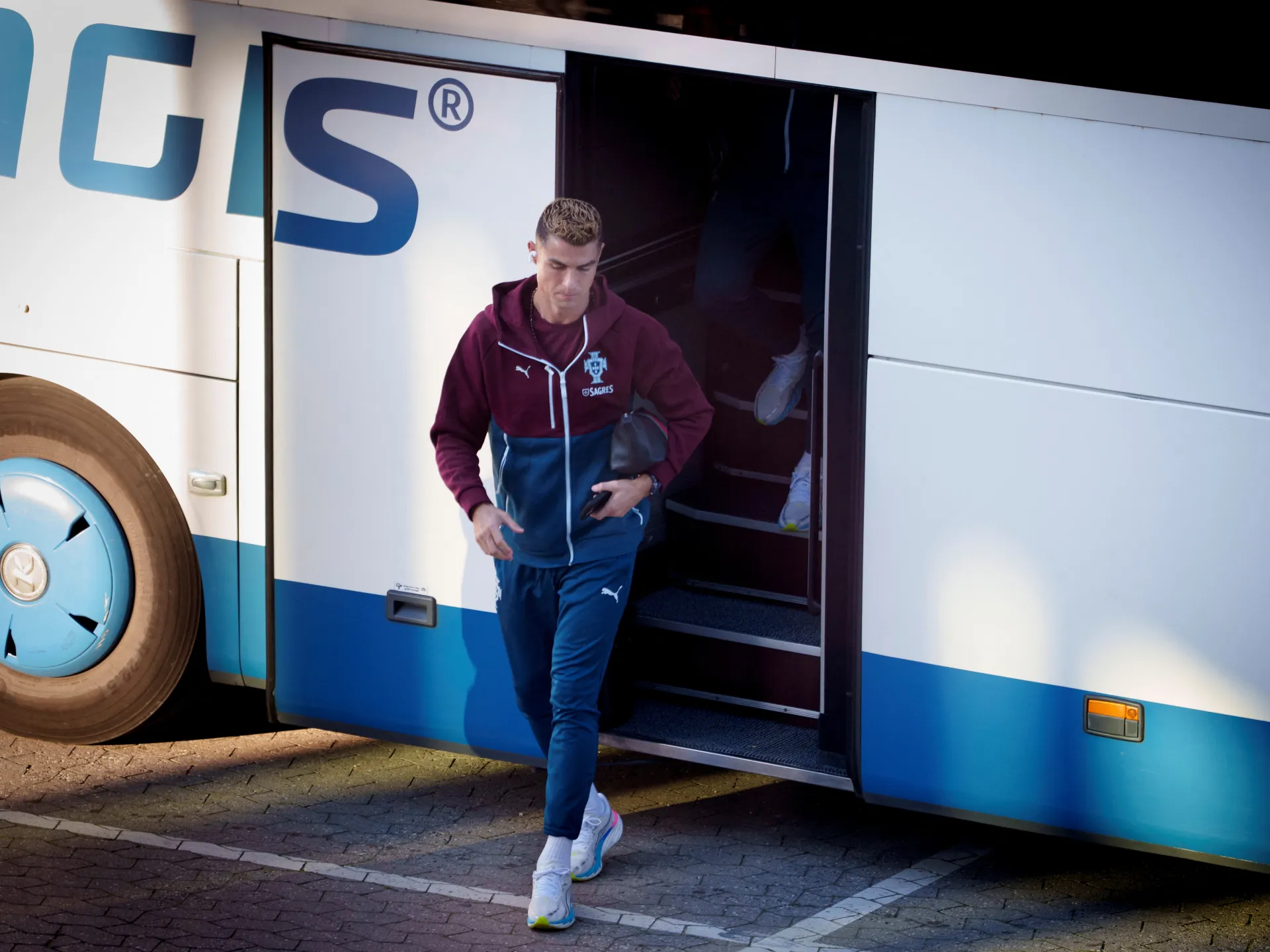 A man in a burgundy and blue tracksuit steps down from a team bus carrying shoes.
