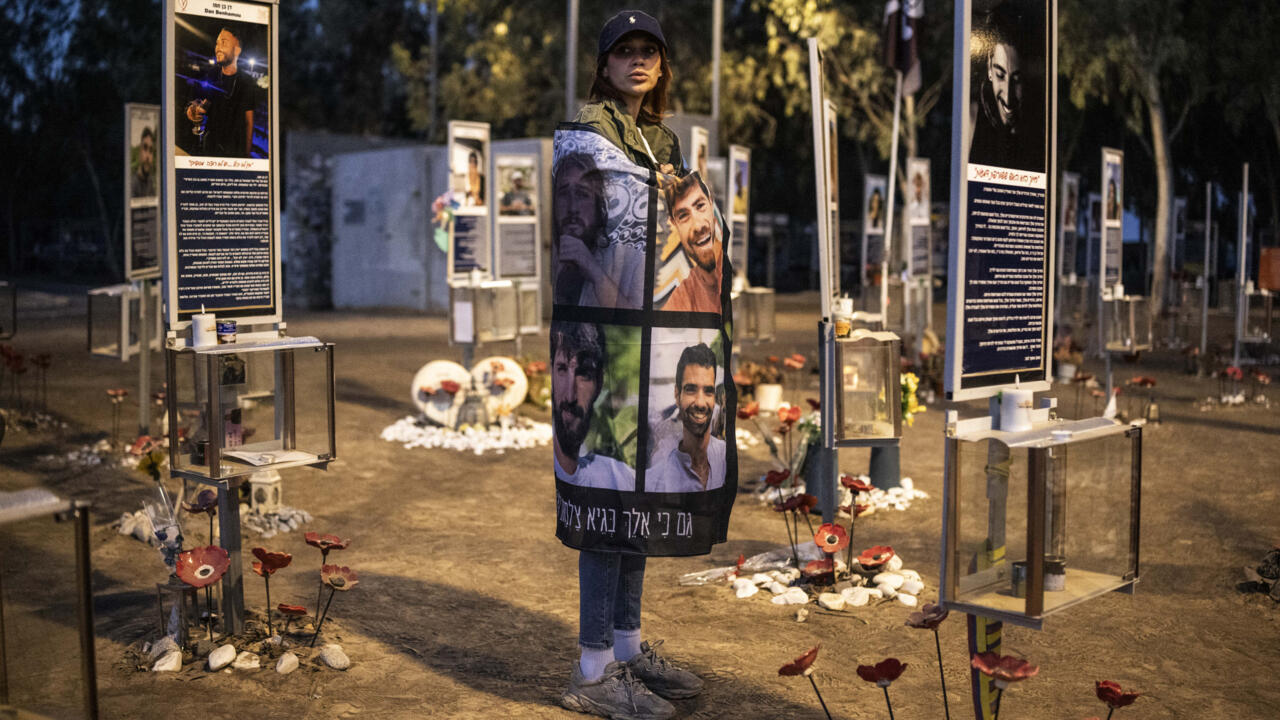 A woman wearing a cap and photo-covered shawl stands among memorial displays, flowers, and candles at night.