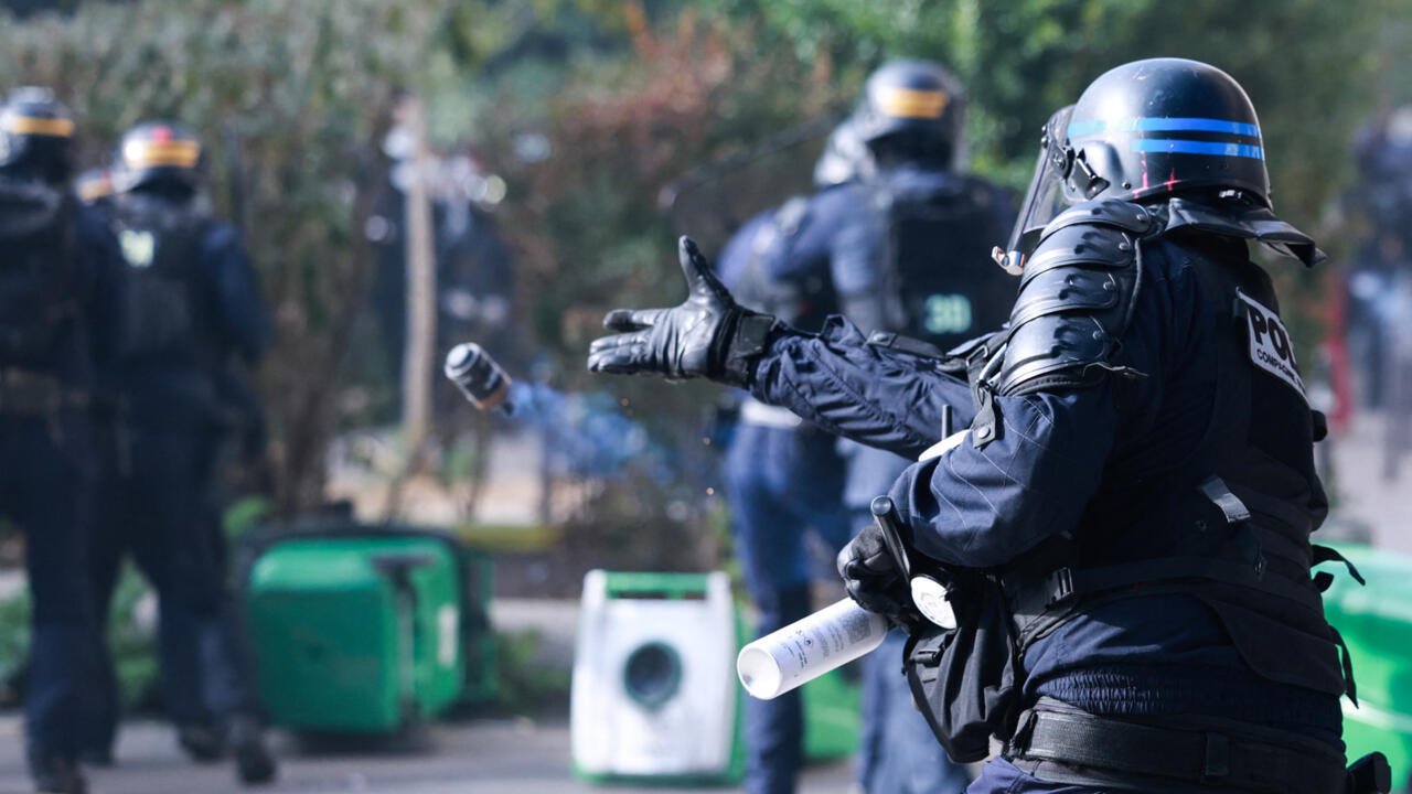 A riot police officer extends an arm while holding a launcher, with other officers blurred behind.