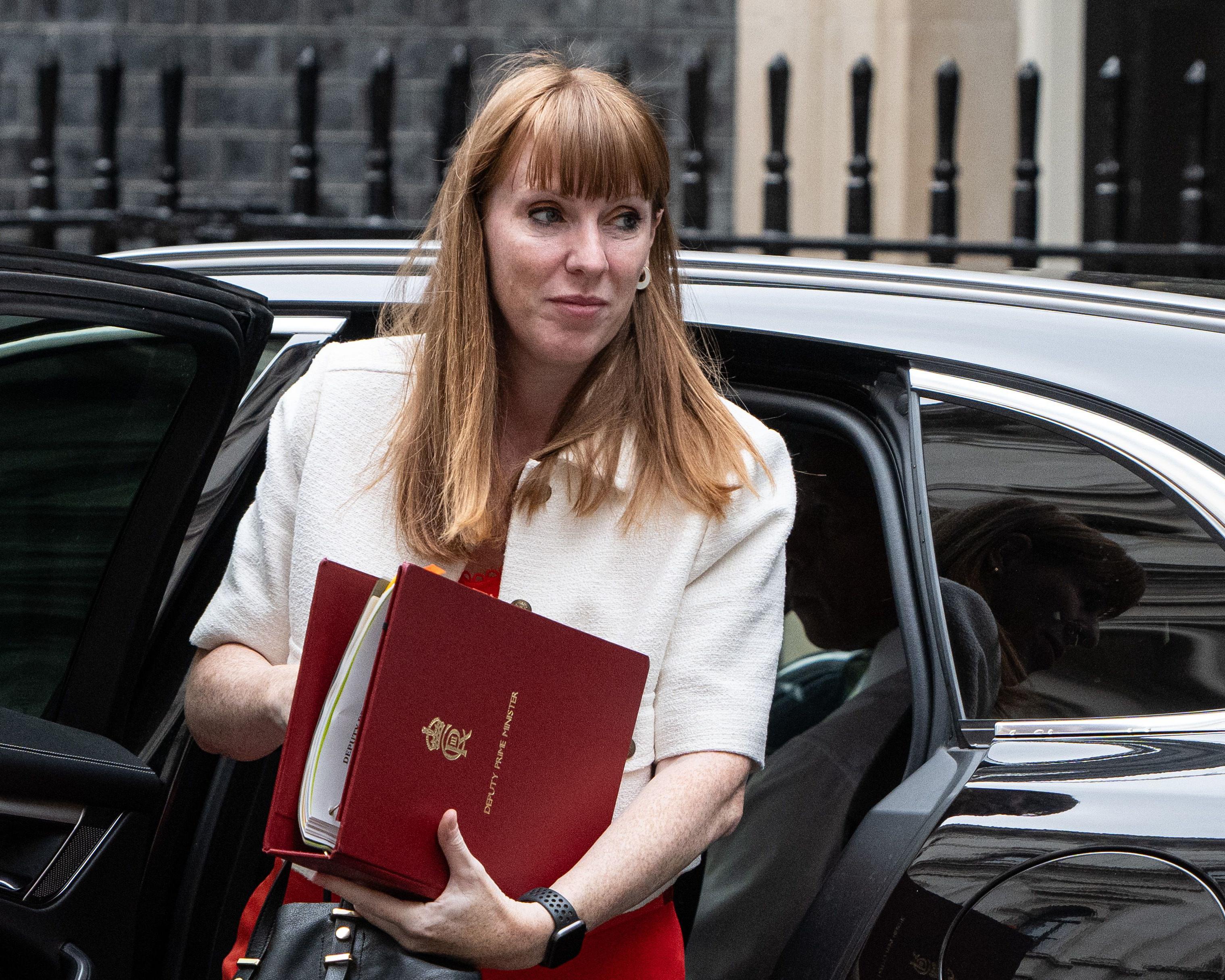 A red-haired woman in a white jacket holds red folders beside an open black car.