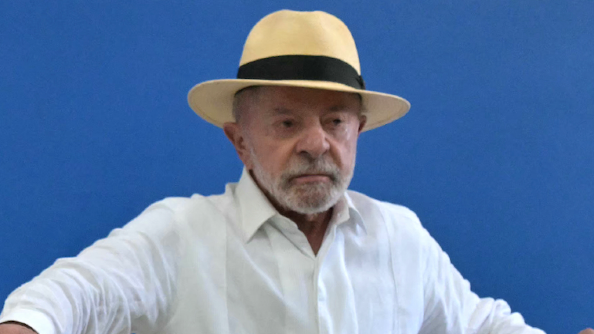 An older bearded man in a white shirt and straw hat sits against a blue background.