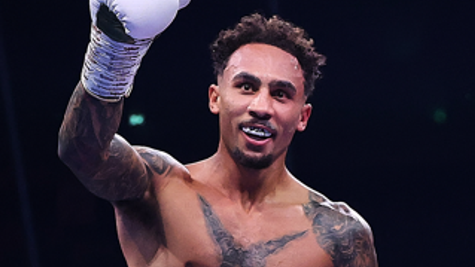 A shirtless boxer with tattoos raises a white-gloved hand against a dark background.