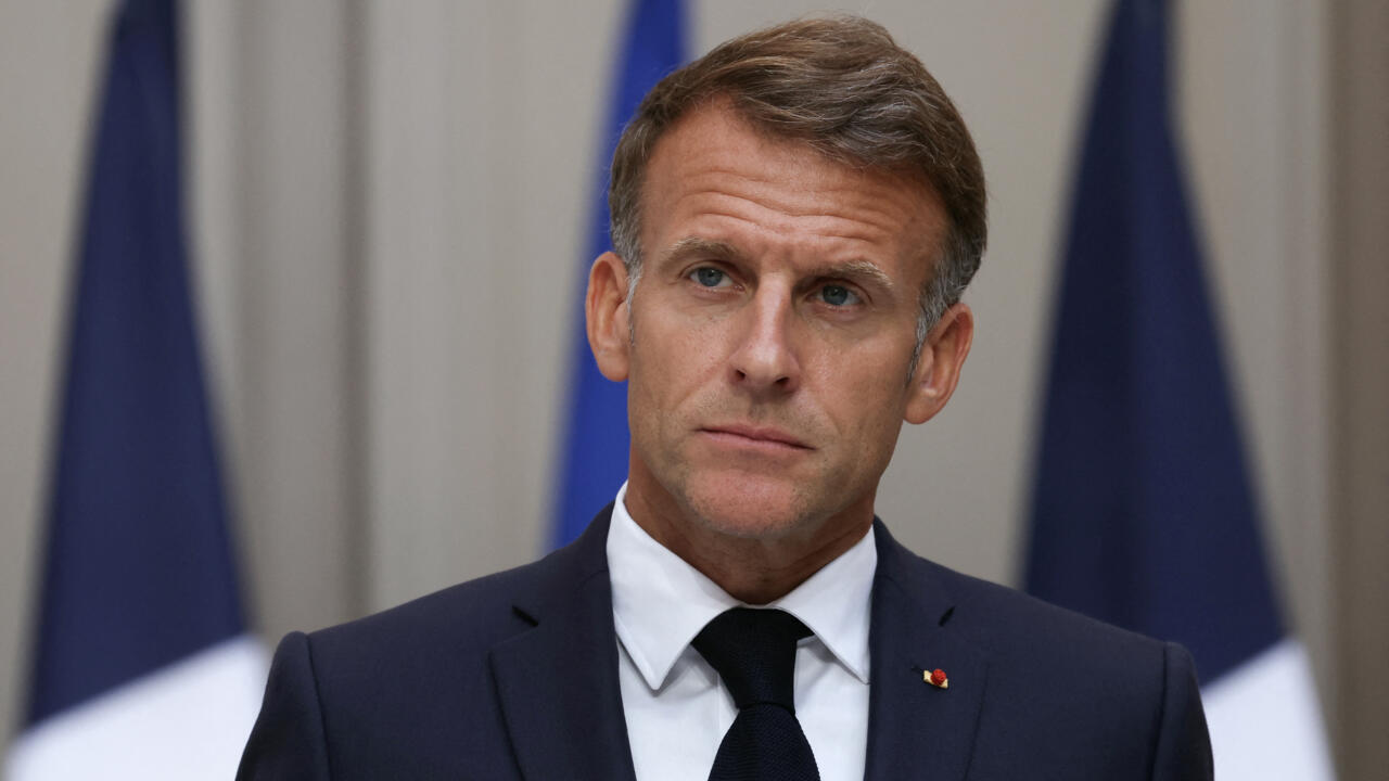 Emmanuel Macron in a dark suit and tie, with French flags behind him.