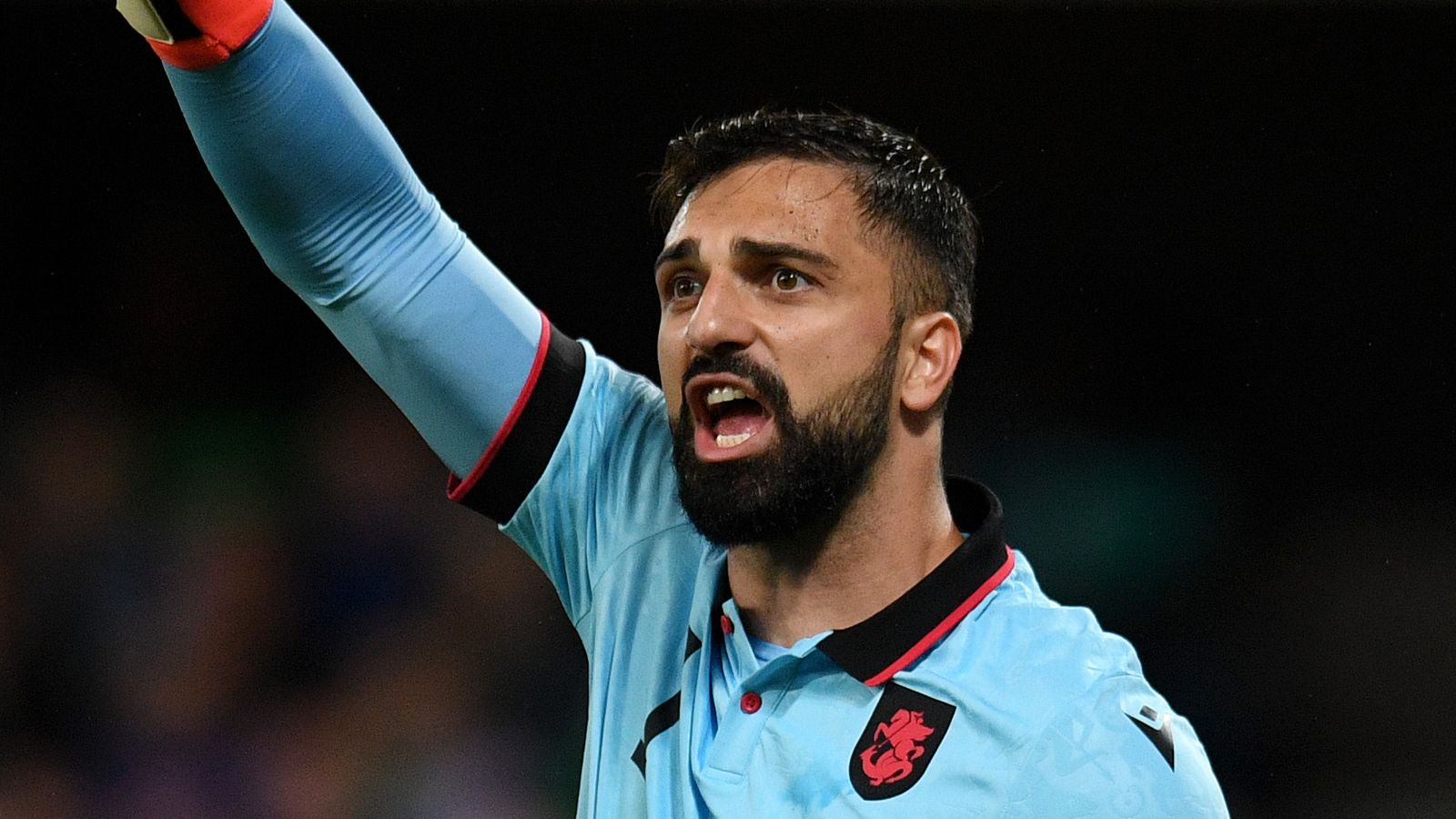 Bearded footballer in a light blue jersey raises an arm and shouts against a dark background.