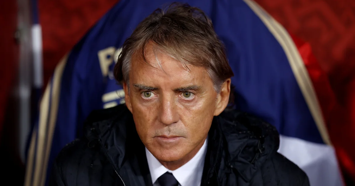Man in a dark jacket and tie sits before a blue, red, and white backdrop.