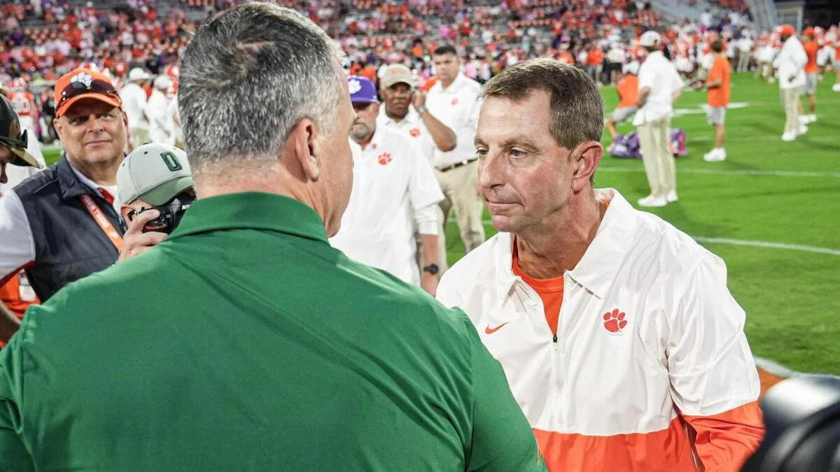 Two coaches face each other on a football field amid players, staff, and spectators.