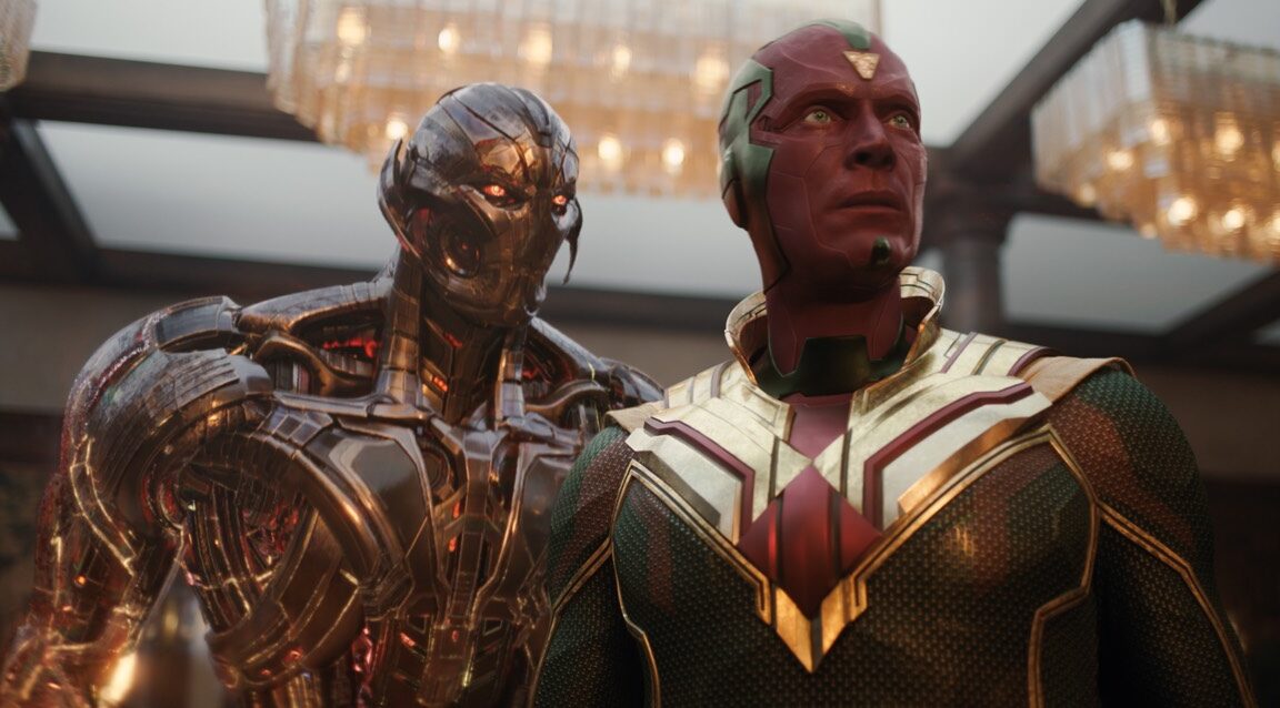A red-faced armored figure stands in front of a reflective metal android beneath warm chandelier lights.