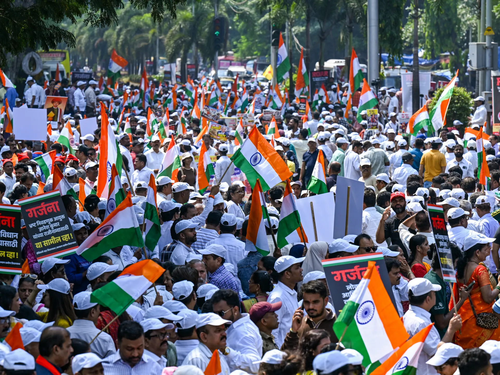 A dense street crowd waves Indian flags and holds placards beneath a tree-lined roadway.