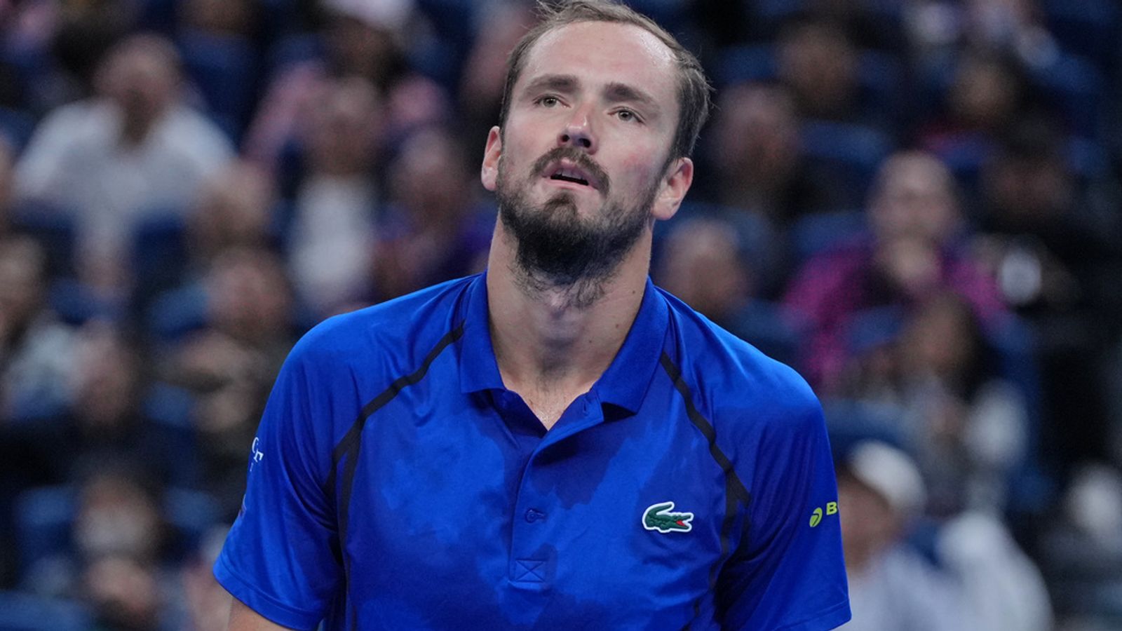A tennis player in a blue shirt looks upward during a match, with spectators blurred behind him.