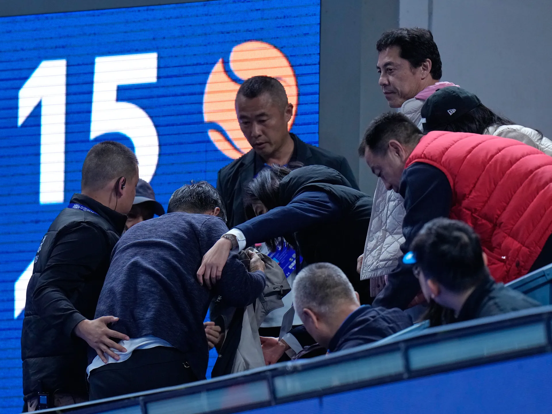 Several men gather around a person bent forward beside blue arena seating and a digital scoreboard.