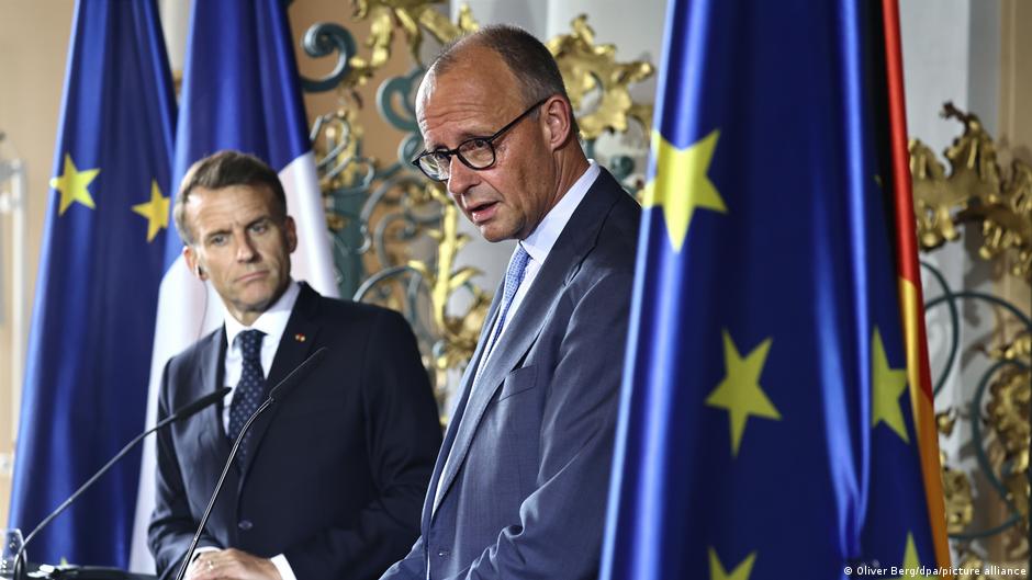 Two suited men stand at microphones amid French and European Union flags.