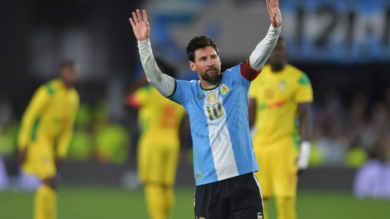 Lionel Messi raises both arms in Argentina's striped jersey as yellow-clad players stand blurred behind him.