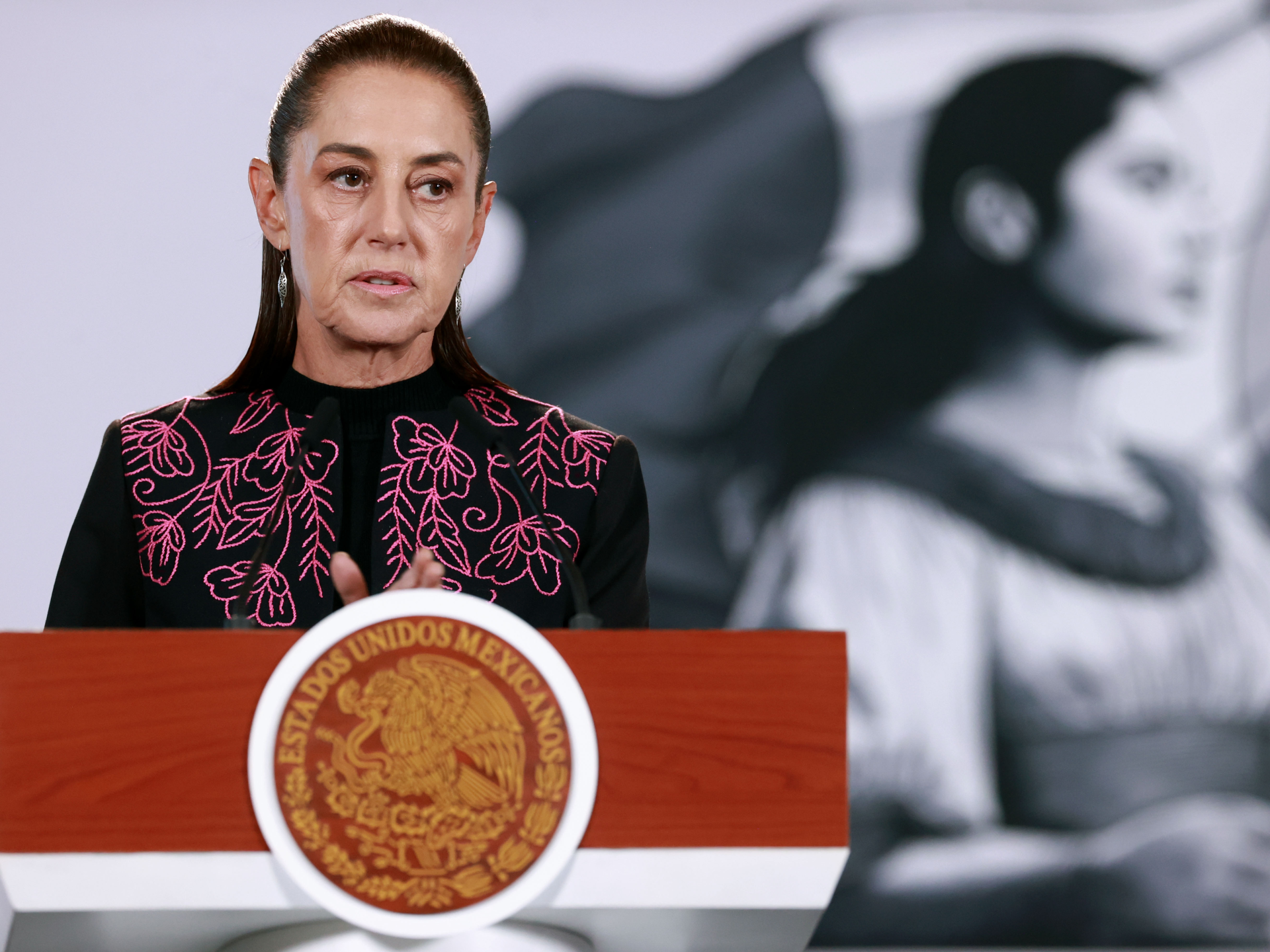 A woman speaks at a wooden podium bearing Mexico’s national seal, with a large black-and-white portrait behind her.