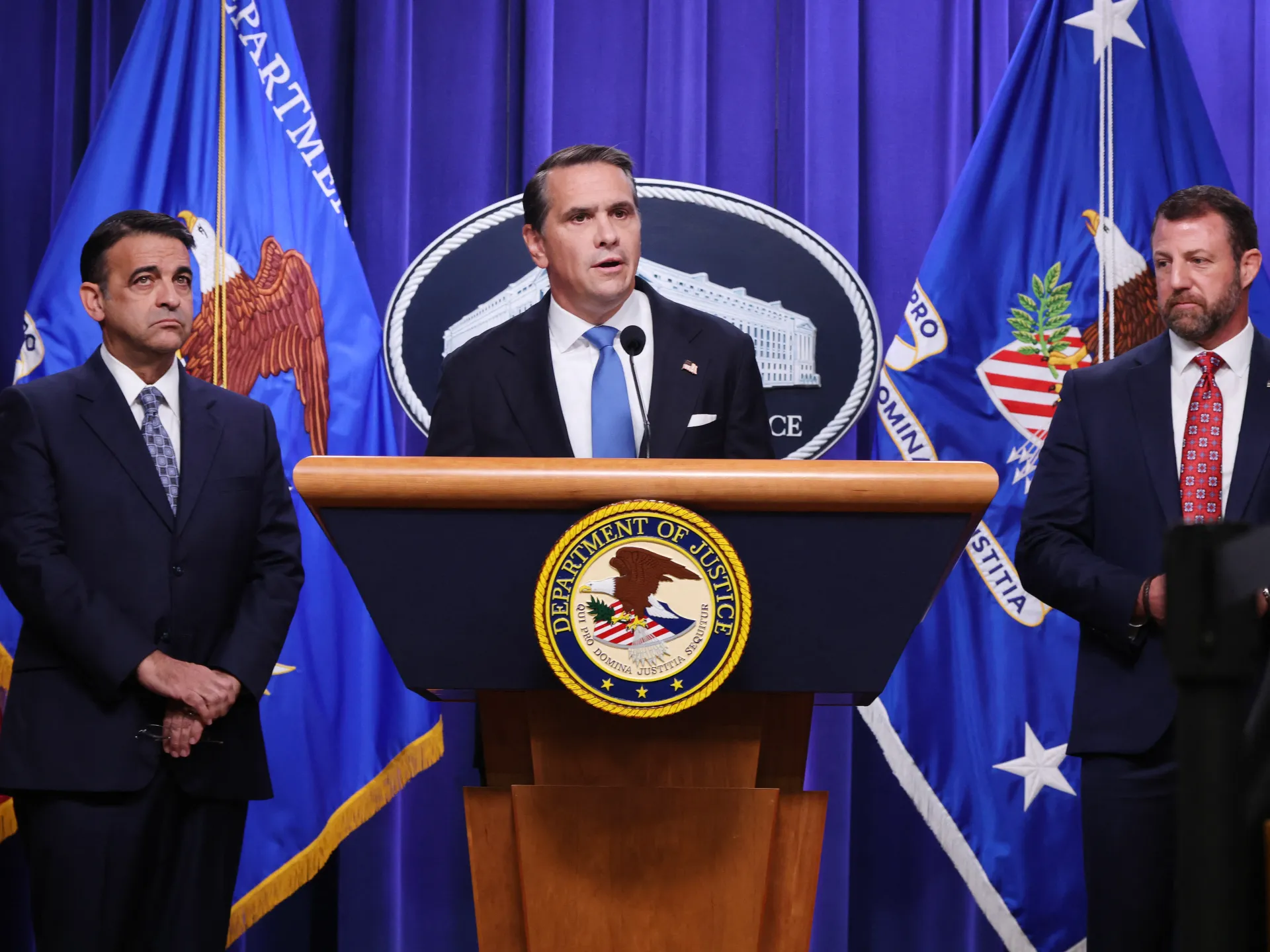 A man speaks at a Justice Department podium between two suited men, with agency flags behind them.