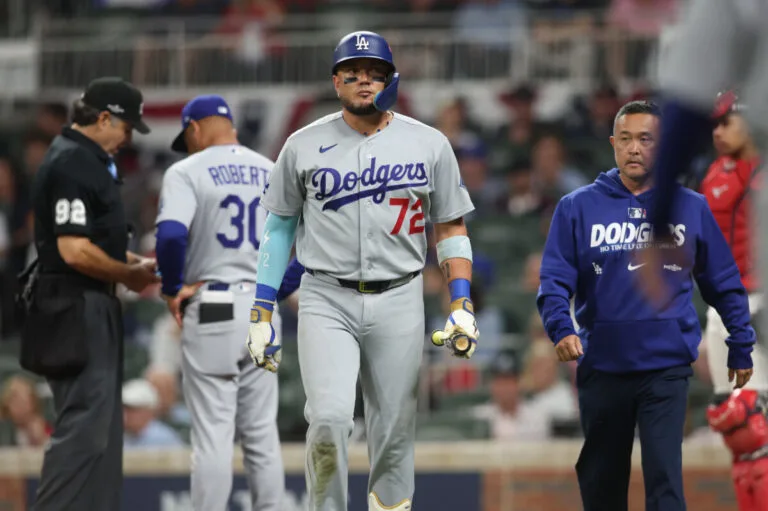 Dodgers player in gray uniform walks beside a staff member as others gather nearby.