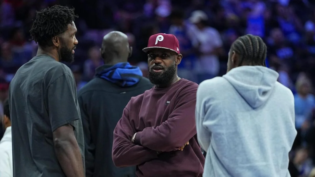Several men talk courtside, including one wearing a Philadelphia Phillies cap and a man in a gray hoodie.