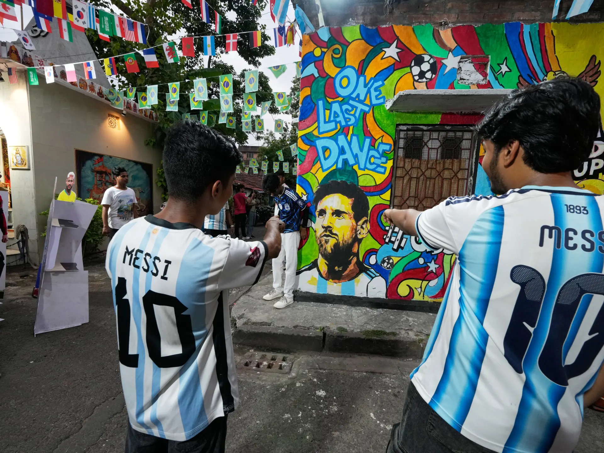 Fans in Argentina jerseys point toward a colorful Messi mural beneath strings of international flags.